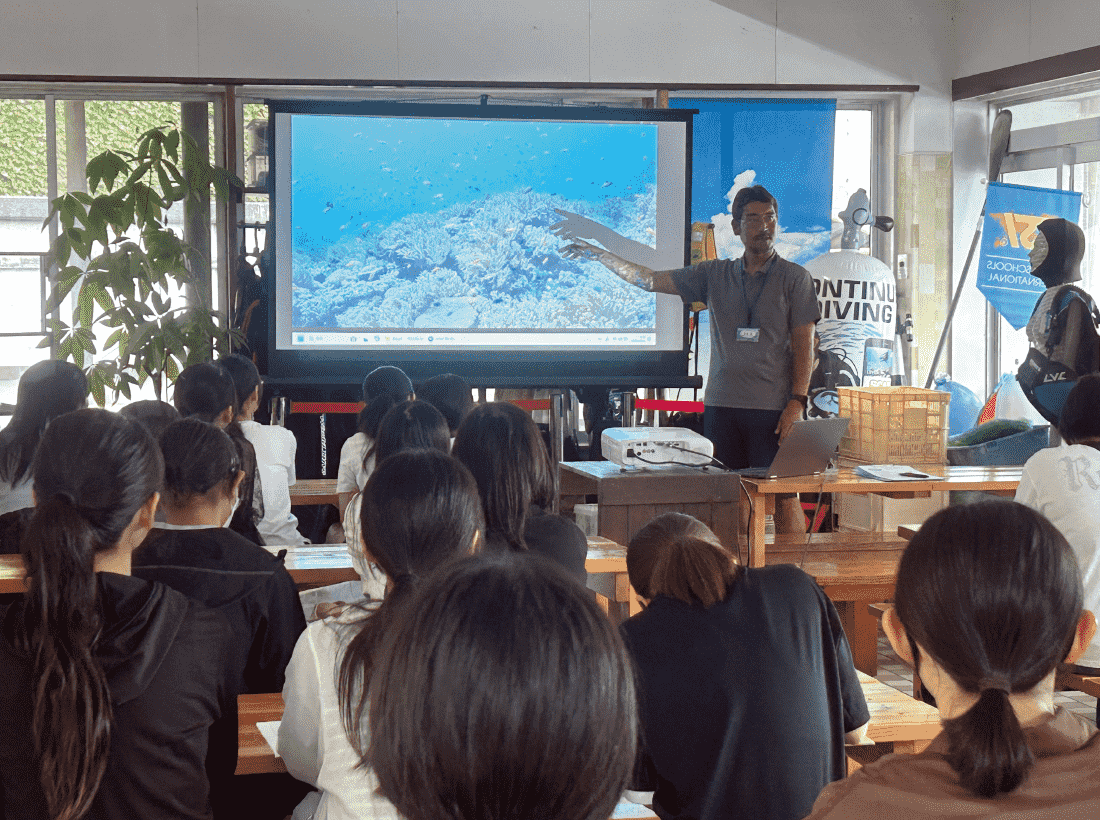愛南町の海と他との海の違いを理解する為の講習中に、西海観光船の館内でプロジェクターを使って海の映像を高校生たちに見せながら説明している